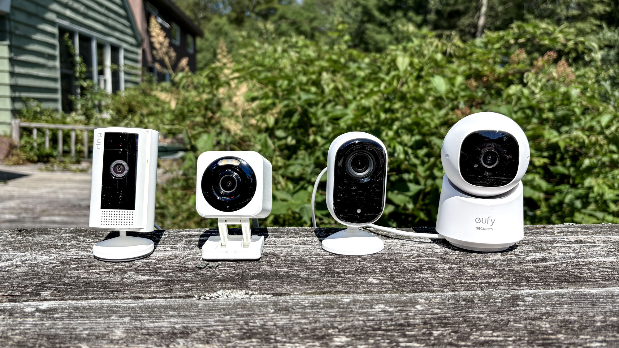 Four home security cameras on an outdoor bench with green bushes in the background