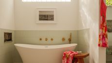 A bright bathroom with a free-standing tub, a wood stool with a colorful towel, a hanging slip dress, a framed painting, a pale green tiled backdrop