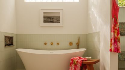 A bright bathroom with a free-standing tub, a wood stool with a colorful towel, a hanging slip dress, a framed painting, a pale green tiled backdrop