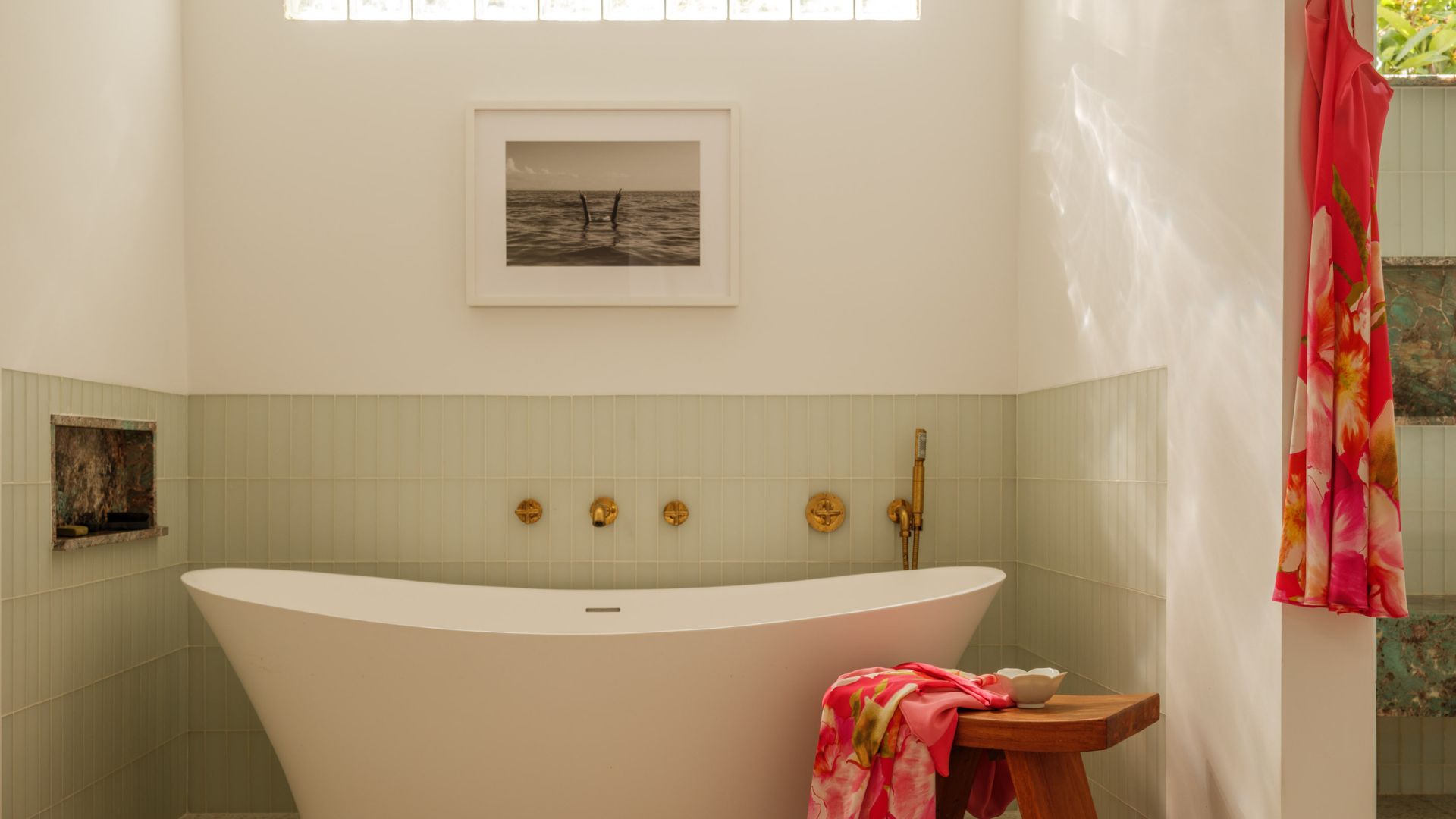 A bright bathroom with a free-standing tub, a wood stool with a colorful towel, a hanging slip dress, a framed painting, a pale green tiled backdrop