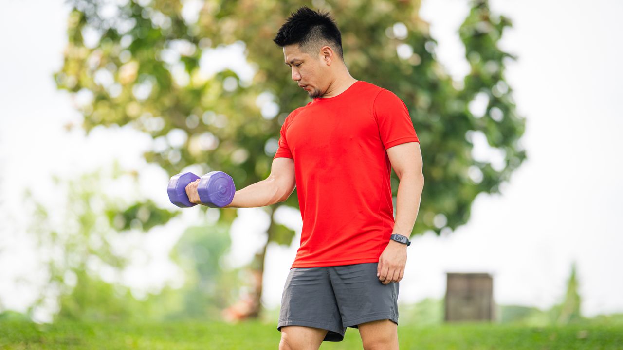 A man in shorts and t-shirts performs biceps curls outside. He is standing, with his left arm by his side and his right arm bent at a right angle as it curls the dumbbell upwards. Behind him we see grass and leafy trees.