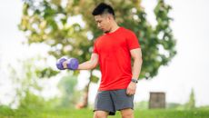 A man in t-shirts and shorts performs biceps curl with a dumbbell outside. He is standing, with his left arm held at his side and his right arm bent at a right angle as it curls the dumbbell upwards. Behind him we see grass and leafy trees.