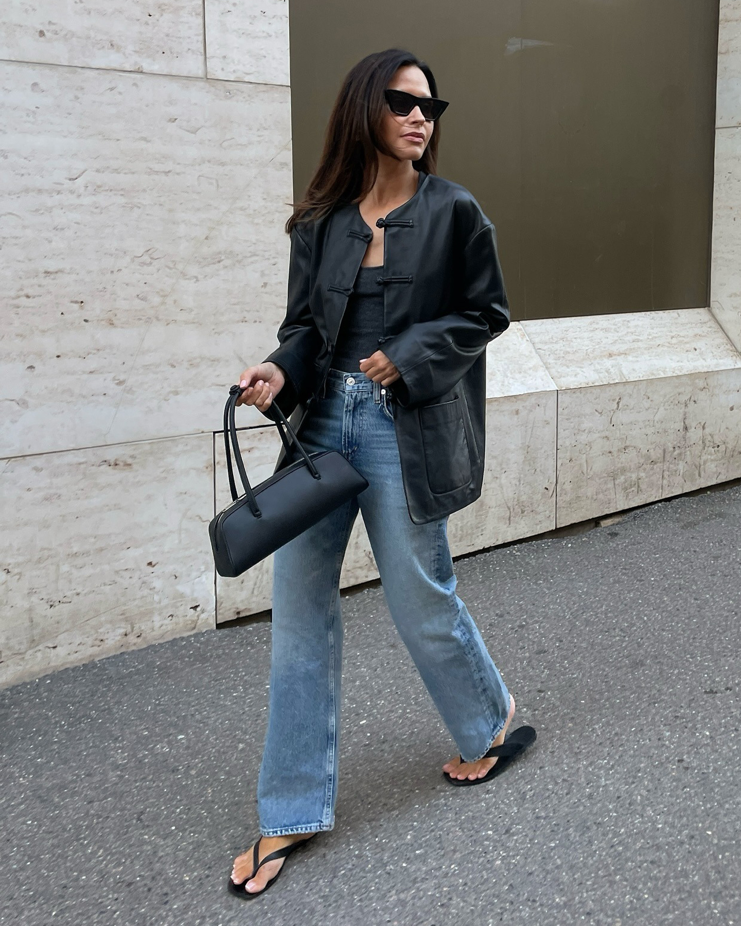 Finnish style influencer Johanna Piispa walking on a sidewalk in Helsinki wearing black sunglasses, a black leather jacket, basic tee, black east-west bag, bootcut jeans, and black flip-flop sandals.