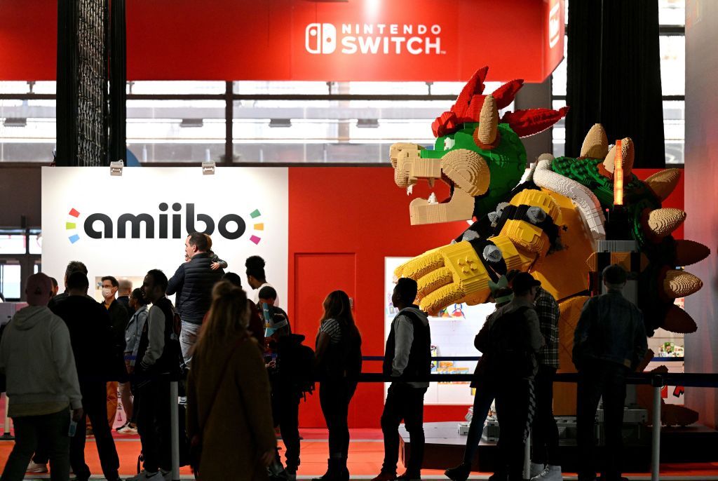 A Lego statue of Bowser from the Mario franchise looms over guests in a convention hall, with Bowser on the right of the frame and guests on the left. Behind them, a large sign reads &#039;amiibo&#039;, and a bright red wall carries the Nintendo Switch logo in white.