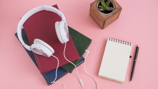Lydbøger: Et par hvide hovedtelefoner ligger ovenpå tre bøger ved siden af en notesblok og en blomst på et lyserødt bord