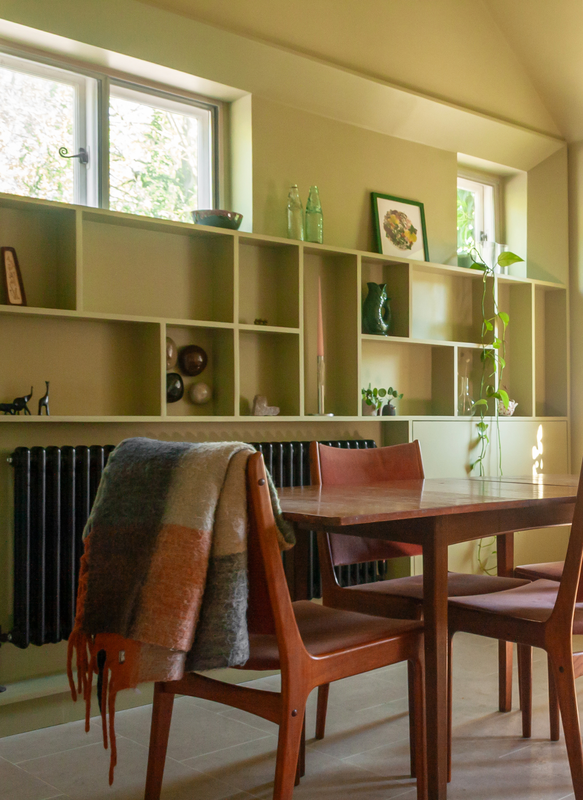 A view of the dining area with colour-drenched walls in a sage/avocado green, showing the wall shelves along the length of the far wall, with the wood table in the middle.