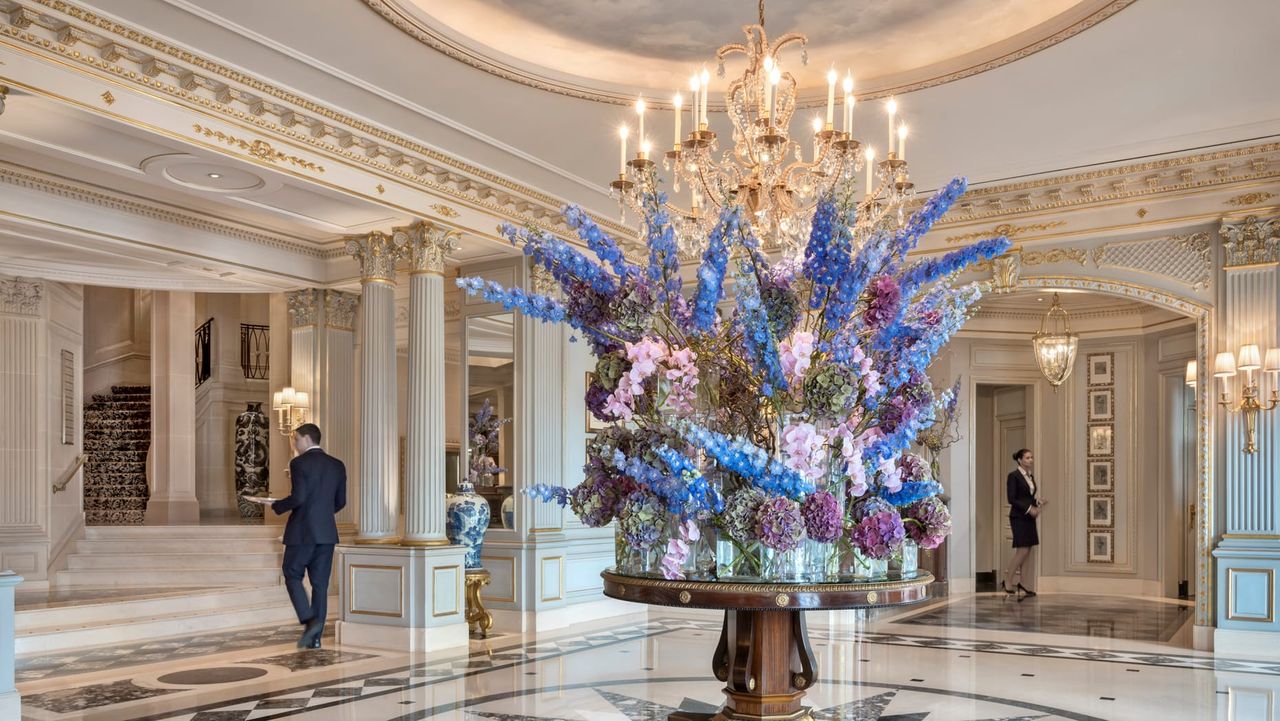 Image of the lobby at Four Seasons Geneva. The walls are white with gold crown molding. There is a large round table in the center with a huge purple flower piece.