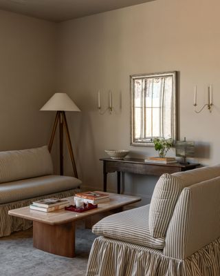 A beige living room with two armless love seats that are facing across from one another. There is a natural wood coffee table between them. In the corner is a wood floor lamp with a white fabric shade.