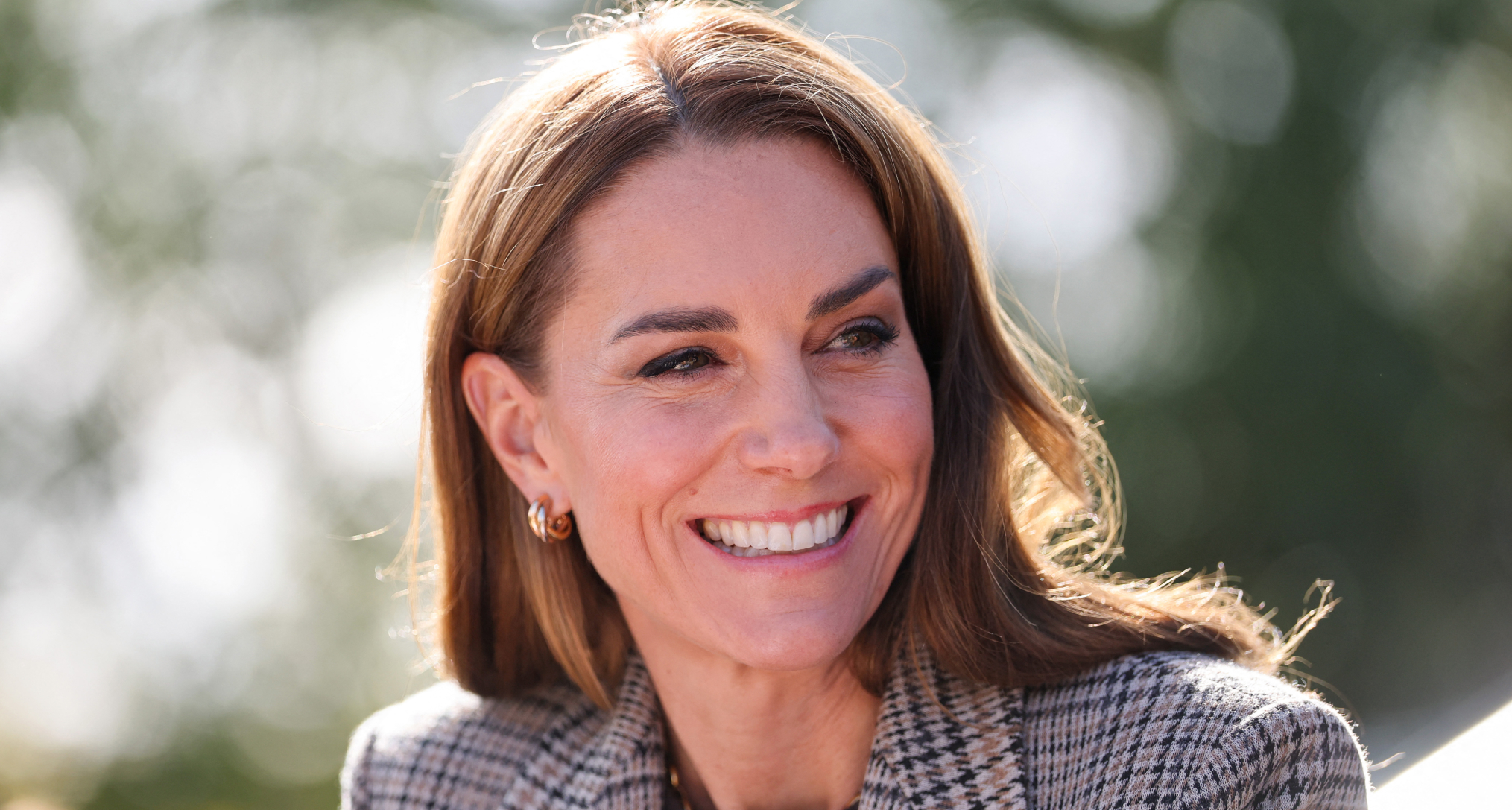 Princess Kate wearing a plaid blazer and smiling in front of a tree