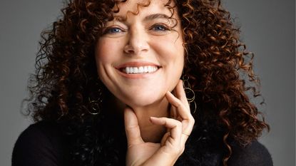A woman with curly hair, golden earrings, and blue eyes smiles at the camera while touching her neck with her left hand.