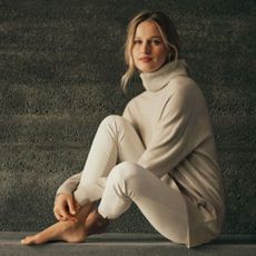 Blonde woman sitting down in cashmere jumper and light jeans with concrete grey background