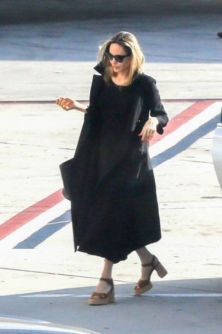 Angelina Jolie steps off a plane in a black coat and a black dress with tall sandals