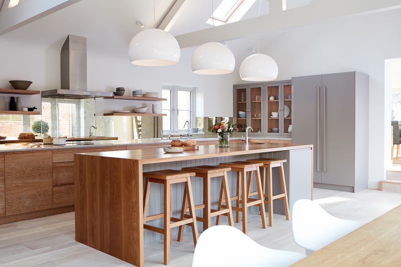 wood and white kitchen with open plan shelving, mirrored backsplash, kitchen island with breakfast bar and stools, large white pendants above, cabinets at one end, white washed flooring, dining table in foreground 