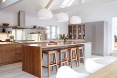 wood and white kitchen with open plan shelving, mirrored backsplash, kitchen island with breakfast bar and stools, large white pendants above, cabinets at one end, white washed flooring, dining table in foreground 