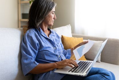 Mature businesswoman sitting on sofa using laptop and analyzing printed charts, working remotely from home