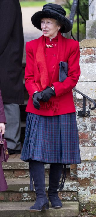 Princess Anne wearing a red jacket and blue plaid skirt with black boots and a wide black hat standing on a staircase outside a church