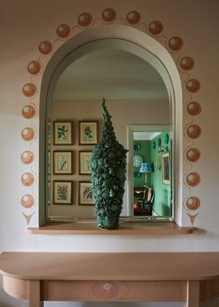 Image of a light pink wall with a window inside a home. The window has a ball trim around it in a more dusty coral shade of pink. There is a green plant in the center of the window