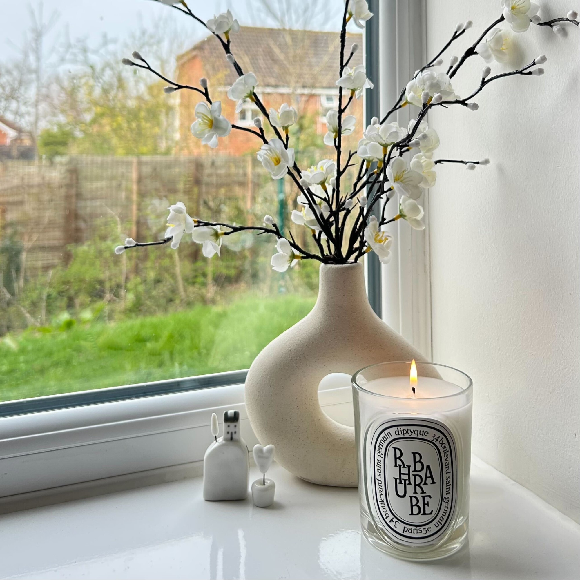 A windowsill at Maddie Balcombe's home displaying Diptyque's Rhubarbe lit candle next to a sculptural cream vase filled with faux flowers