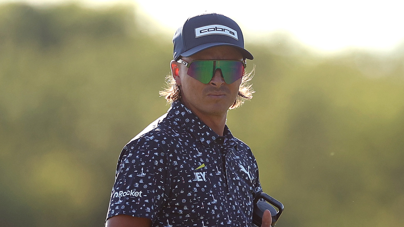 Rickie Fowler from the chest up wearing a navy polo, navy cap and sunglasses at the US Open
