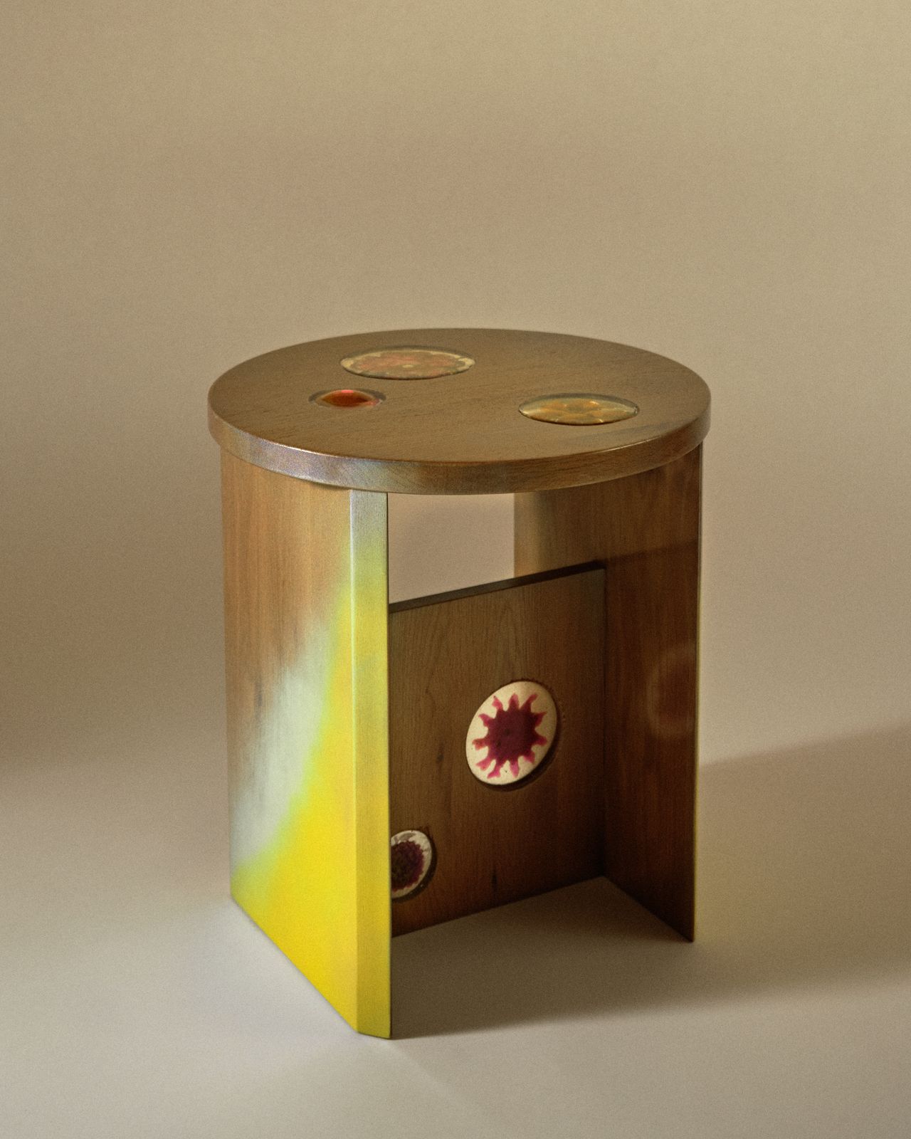 A fanciful envisioned stool features a gradient of yellow, pink, and light blue tones on its wooden surface, as well as sun-shaped motifs.