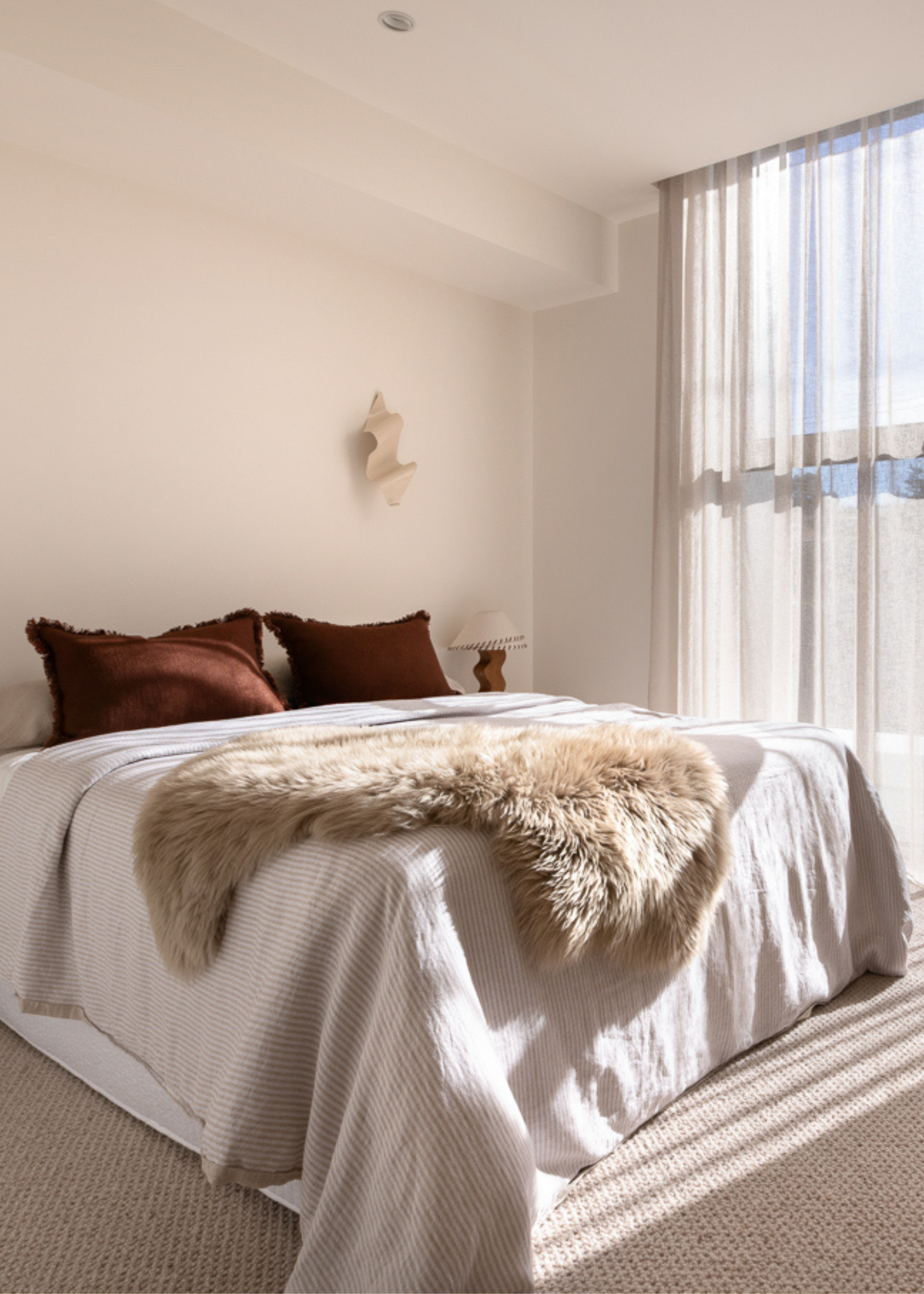 A white bedroom with sheer window treatments, a curved wall sconce, a wood lamp, and a bed with a striped duvet, a fuzzy throw, and a set of brown pillows