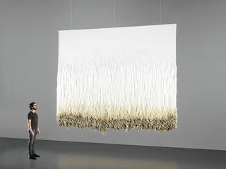 A man in tshirt, jeans and shoes staring at a larg e artistic display of wool hanging from the ceiling depicting the transition of wool from its raw state to refined weave