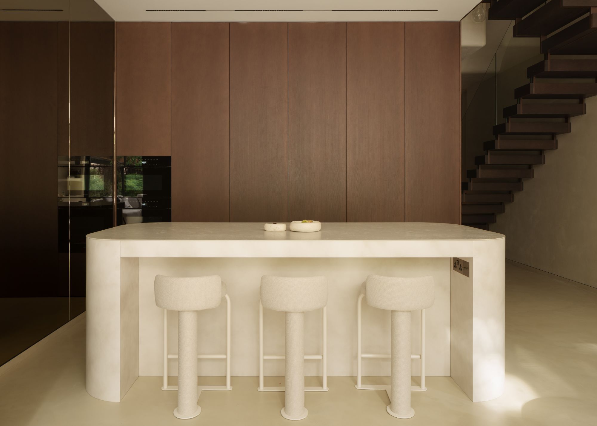 A rounded, cream-colored marble kitchen island with three cream bar stools, in front of a hidden kitchen concealed behind sliding timber doors