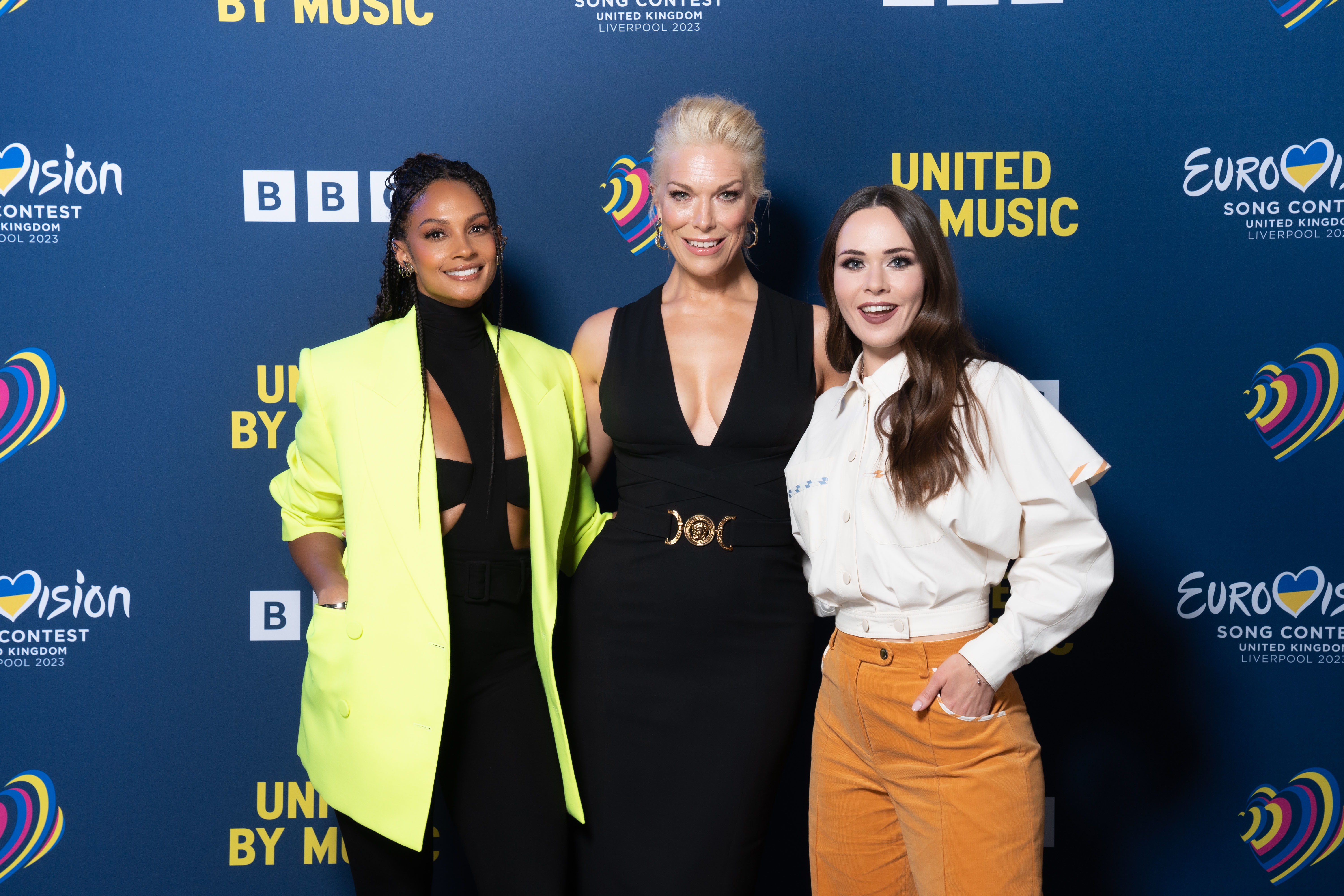 Alesha Dixon, Hannah Waddingham and Julia Sanina posing together at the BBC's official Eurovision Song Contest launch in Liverpool, standing in front of a display board  that shows the BBC logo, the Eurovision Song Contest logo, and the slogan 'United by music'