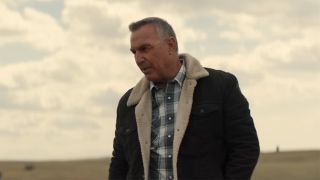 Kevin Costner in a plaid shirt and shearling coat looking down at a field and talking in Let Him Go. 
