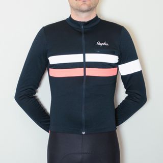 A white man in a dark blue long sleeve cycling jersey and black cycling shorts stands against a white wall