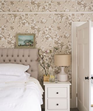 Floral patterned bedroom wallpaper ideas in beige, with white bedroom furniture and a small gold-framed painting resting on the headboard.