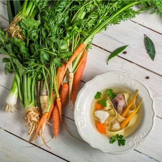 Carrots, celery root, and leeks next to a bowl of vegetable noodle soup