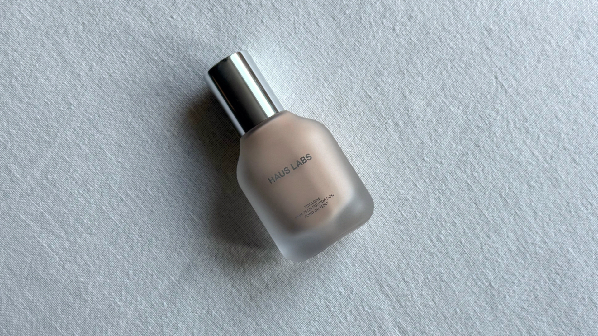 A bottle of the Haus Labs Triclone Foundation laying at an angle against an off-white linen background