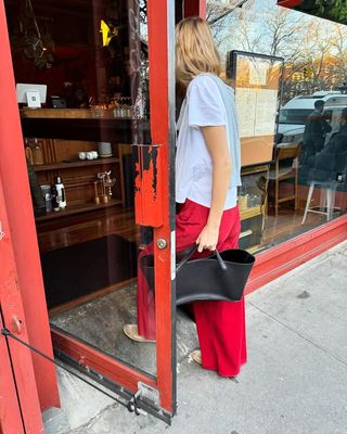 Courtney Grow is wearing red wide-leg pants, mesh flats, a black bag from The Row, and white t-shirt. She also has a sweater slug over her shoulders.