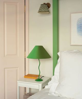 Green table lamp with wiggle base in white bedroom, placed on white bedside table, white bedding, green painted wooden panel