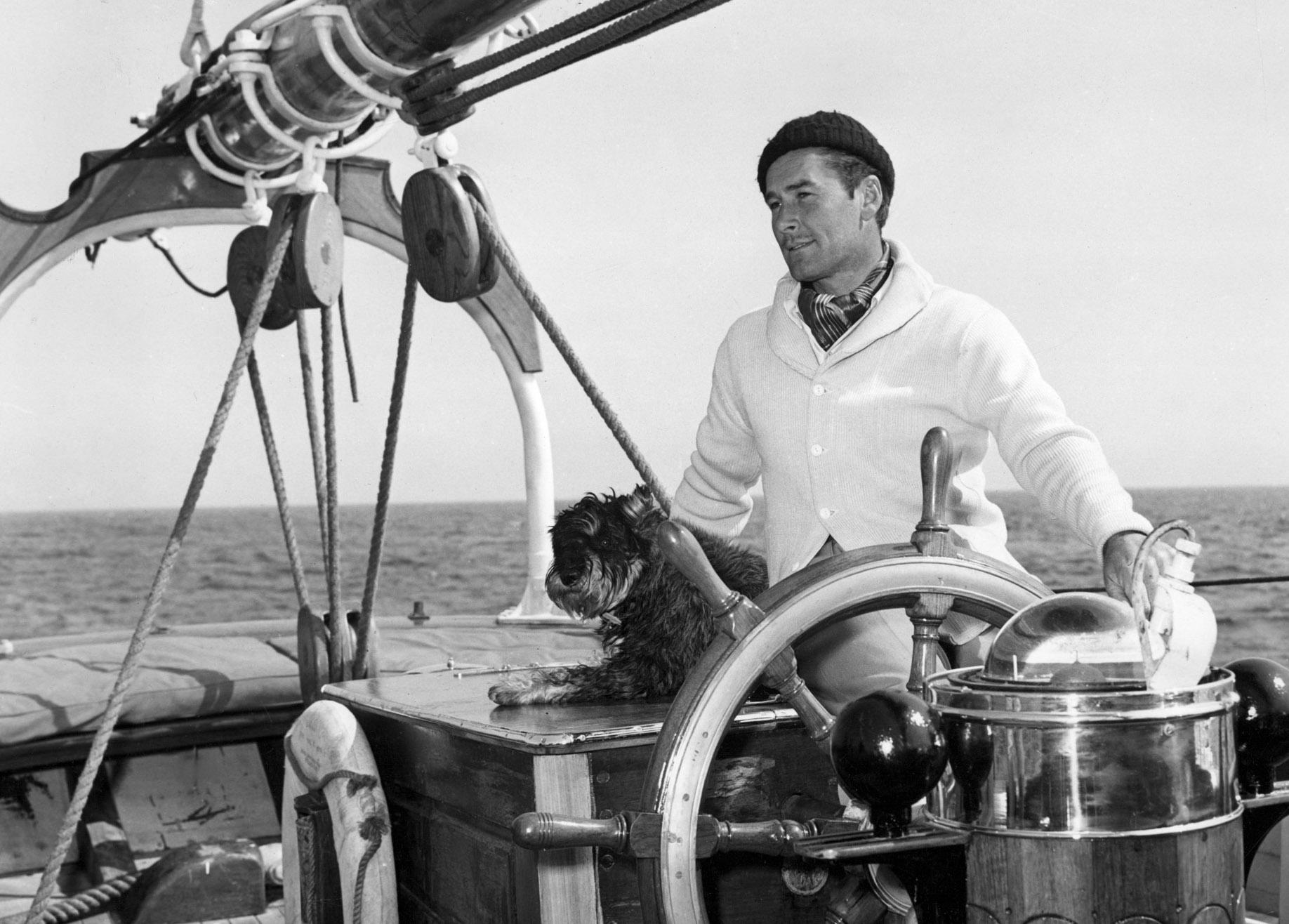 Errol Flynn behind the wheel of a sailing yacht with a small dog