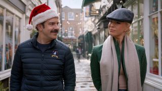 Ted in a Santa hat and Rebecca in a paperboy hat, they're walking down the street smiling at each other.