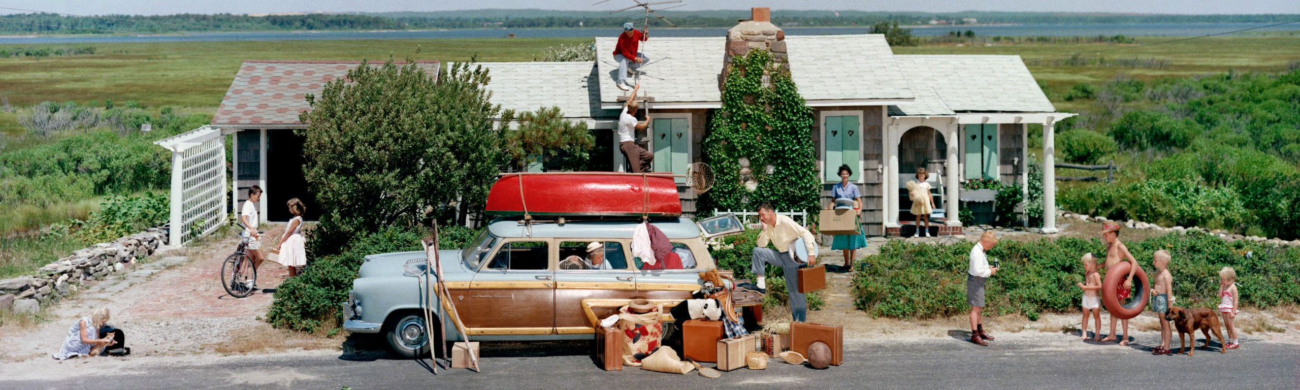 A 1950s family pack up outside a shingled cottage, a red canoe strapped to the roof of a station wagon, suitcases piled on the road and children, a dog and a man fixing a TV aerial on the roof.