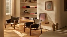 Image of a beige living room with a large built-in bookshelf with several stylish coffee table books stacked on it. There are two black leather and wooden accent chairs with a small white marble side table between them