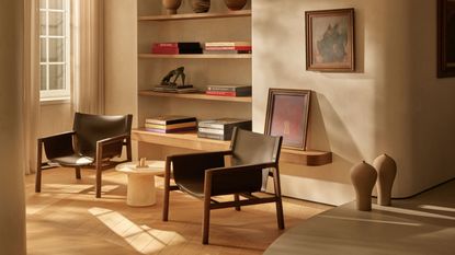 Image of a beige living room with a large built-in bookshelf with several stylish coffee table books stacked on it. There are two black leather and wooden accent chairs with a small white marble side table between them