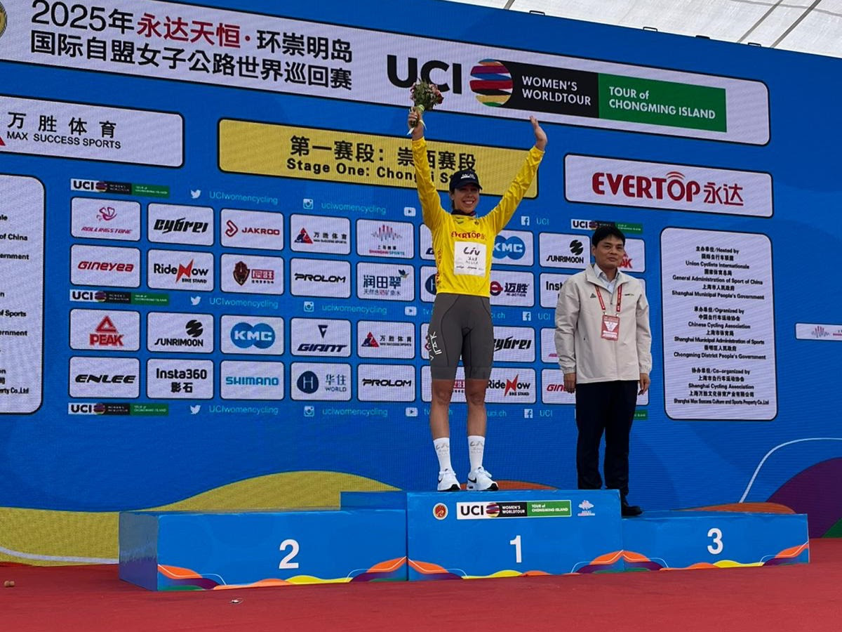 Georgia Baker on the podium at the Tour of Chongming Island, wearing a yellow leader&#039;s jersey and standing with her arms aloft in celebration