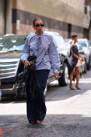 A guest wears black sunglasses, gold earrings, light purple flowy oversized shirt, shiny black leather bag with black tassels, dark blue denim jean baggy pants, shiny red pointed toe heels, outside Tibi, during the New York Fashion week Spring/Summer 2025 on September 07, 2024 in New York, New York.