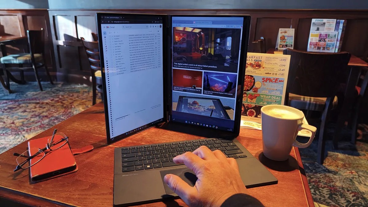 A dual-screen laptop is set up in a vertical orientation with the detachable keyboard in front, and a hand is shown resting on the trackpad.