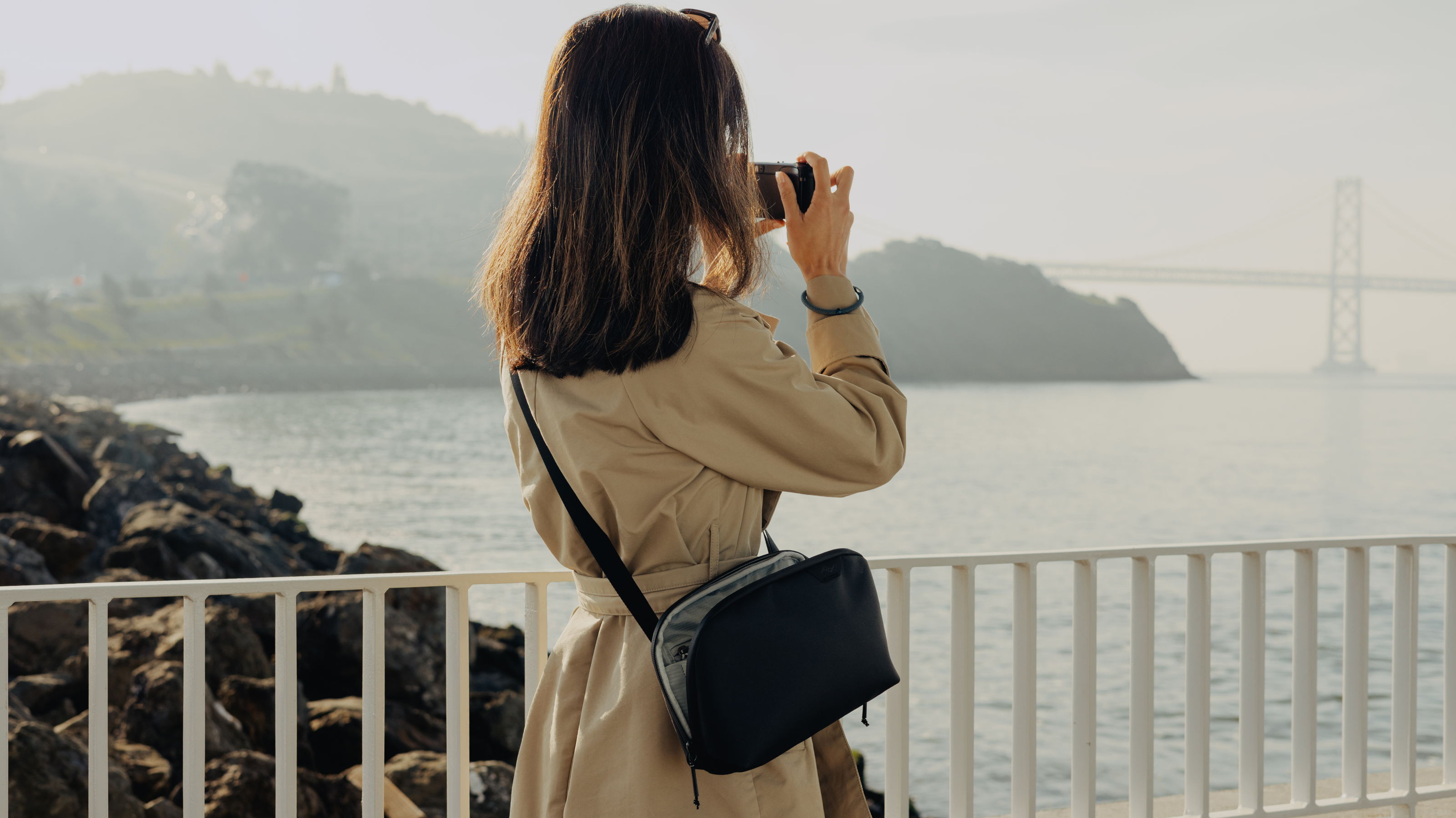 Woman wearing the Peak Design Travel Crossbody 3L bag while taking a photograph of a river and a bridge with a small compact camera