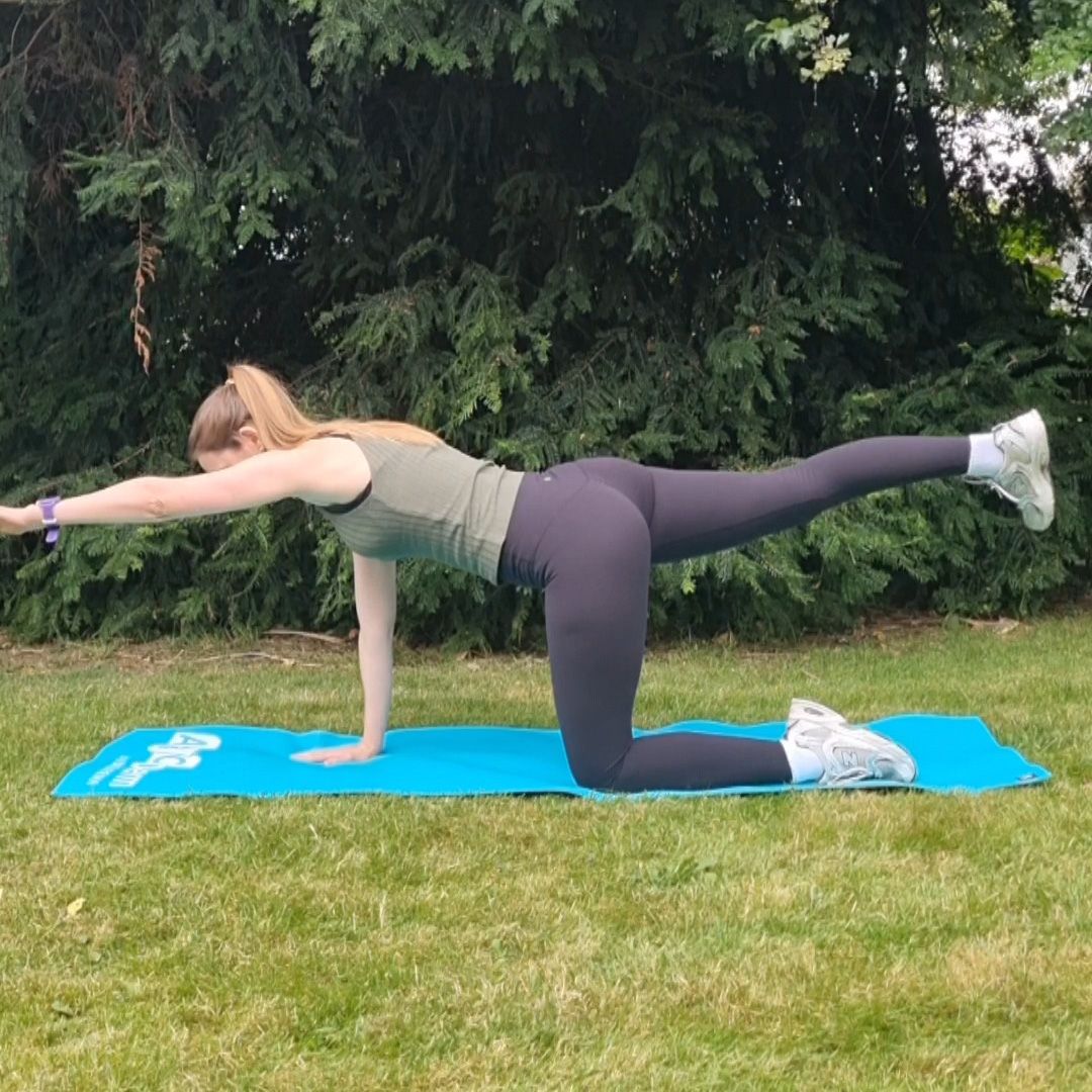 Fitness editor Ruth Gaukrodger performs a bird dog exercise in an outdoor setting on a yoga mat. Her left knee and shin lie on the yoga mat, and her right hand is on the ground, while her right leg and left arm are extended outwards, parallel with the ground. Behind her we see a pine tree.