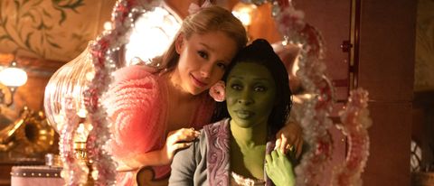 L to R: Ariana Grande is Glinda leaning against Cynthia Erivo is Elphaba as they look into a mirror. 