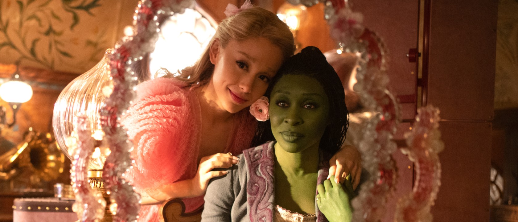 Ariana Grande as Glinda stands beside Cynthia Erivo’s Elphaba, both gazing into a mirror.