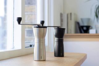Two Hario Mini Slim Pro hand grinders, one in silver and one in black, standing on a kitchen worktop