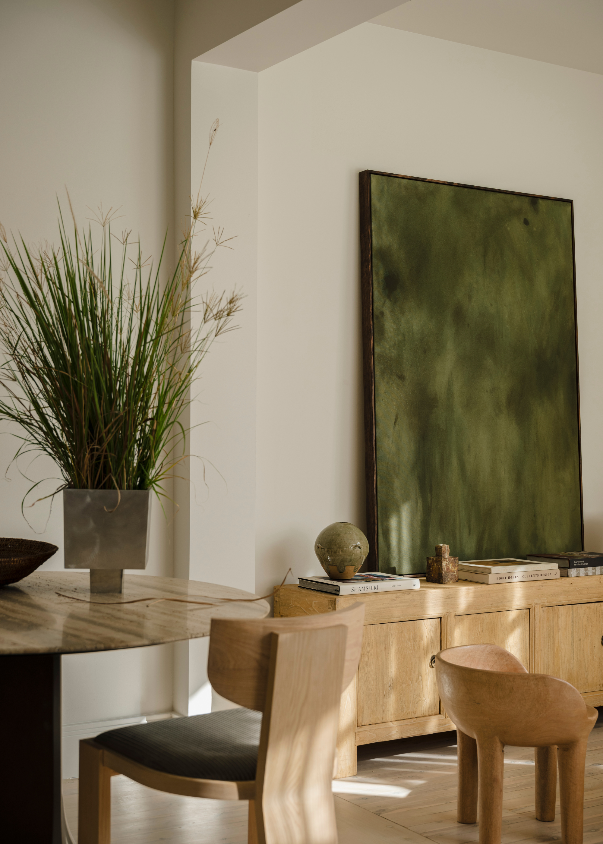 A dining room with a potted plant on the dining table and a sideboard with a green art piece, decor and books