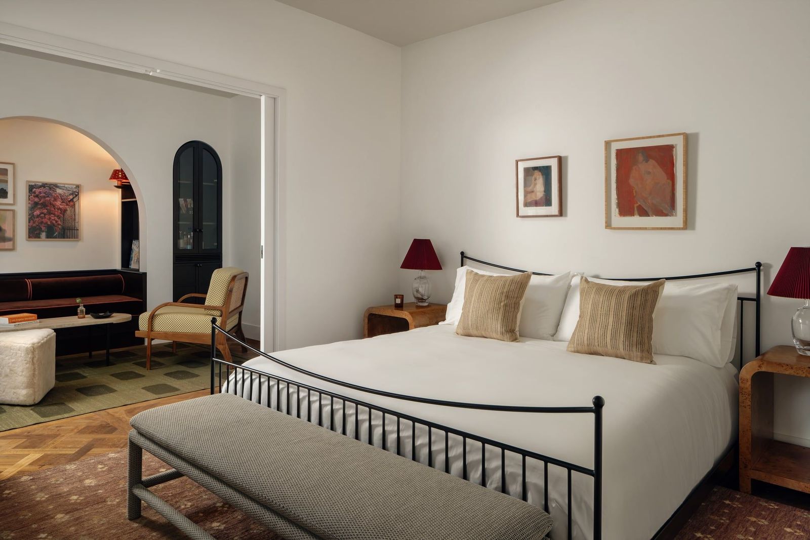 A hotel suite with wooden floors, earthy decor in dark red, olive green, and neutral tones, a wrought iron, essential bedroom, and an arched living area housing a cozy soft seating nook.